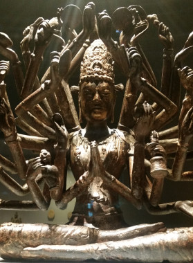Avalokitesvara statue, with many arms, sitting cross-legged in prayer. The bronze sculpture is dramatically lit from the top. The many hands holds symbols of Buddhist ideals, with eyes closed, two front hands held together in prayer. The statue has many detailed patterns.