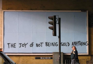 A huge white board on the street behind a street light, a black handwritten sentence written on it, just a bit over the board up until to the frame: "The joy of not being sold anything", all caps. The street sign below the board says MAIDEN, two women passing by.