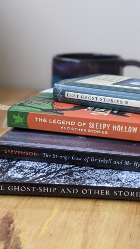 A photograph of a pile of books on a light wooden desk. Visible are Best Ghost Stories, a collection of Washington Irving stories, a collection of Robert Louis Stevenson stories, and a collection of Richard Middleton stories. Blurred in the background is a cool coffee mug.