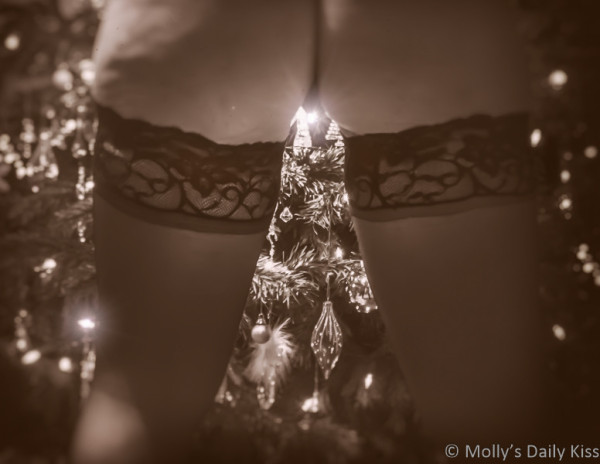 Molly standing with legs apart in front of Christmas tree wering hold up stocking. We can see her bare bum, back of her legs and stocking tops, through her legs we can see the Christmas tree with ornaments and lights. Once of the light is shining bright right by her vulva lips. The whole image is edited in a sepia tone