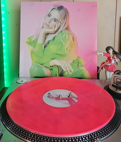 A Pink [Fluorescent] vinyl record sits on a turntable. Behind the turntable, a vinyl album outer sleeve is displayed. The front cover shows Laurel sitting and resting her cheek on her hand. 

To the right of the album cover is an anime figure of Yuki Morikawa singing in to a microphone and holding her arm out. 