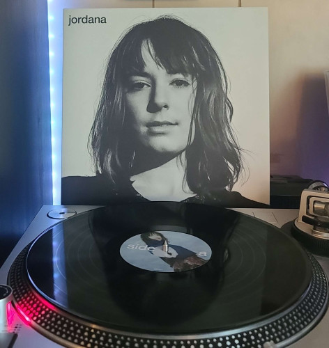A black vinyl record sits on a turntable. Behind the turntable, a vinyl album outer sleeve is displayed. The front cover shows a face shot of Jordana in black and white
