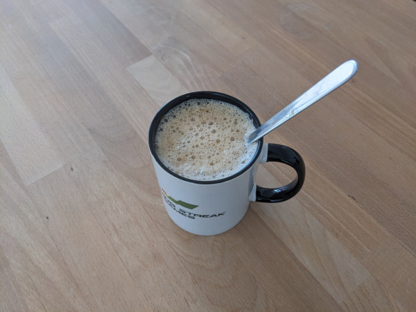 A cup of coffee resting on a wooden table. The handle of a tablespoon is hanging out of the cup.