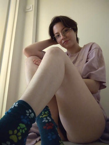 Poki (slim white person with short dark hair and big prescription glasses) sitting on the floor, wearing dark green socks with paws printed on them, no pants (but black panties) and a light purple oversized tee. The camera is placed so their naked thighs (one mostly) take the most of the shot. They're softly looking and smiling at you