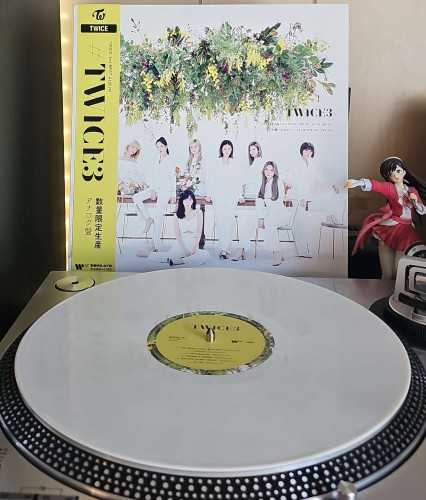 A white vinyl record sits on a turntable. Behind the turntable, a vinyl album outer sleeve is displayed. The front cover shows the 9 members of TWICE sitting on chairs and desks while wearing white suits. A large plant is hanging above them . 

To the right of the album cover is an anime figure of Yuki Morikawa singing in to a microphone and holding her arm out.