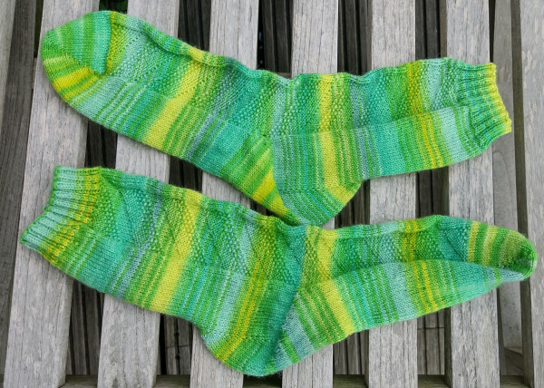 A pair of handknit socks on a background of weathered wood. Socks are in different shades of bright greens in a striped pattern. There's a bit of textured pattern on the socks.
