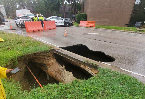 A large hole  on the left side of a city street, with most of the hole in the grass.