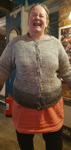 Laughing fat woman with cane proudly wearing hand-knit cardigan with descending gradient of colour from cream to darkest brown.