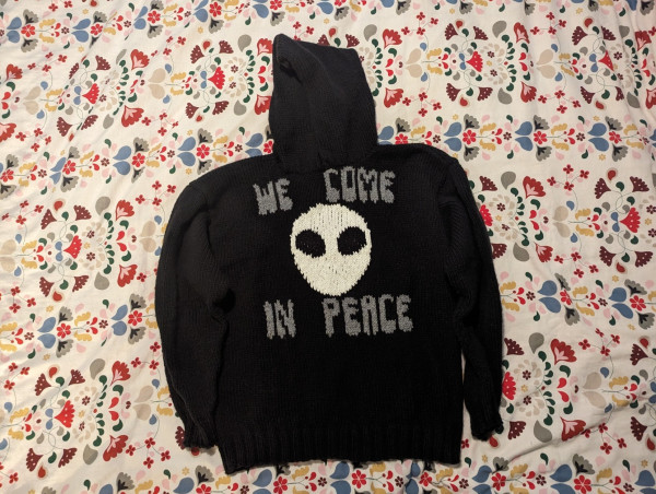 Hand knit hoodie with colour work on the front saying, "WE COME IN PEACE", with a "grey" space alien face in the centre.