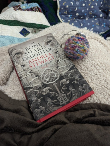 A knitted bauble and a book sit on a pile of quilts and other blankets. The book is The Bone Share Daughter by Andrea Stewart.