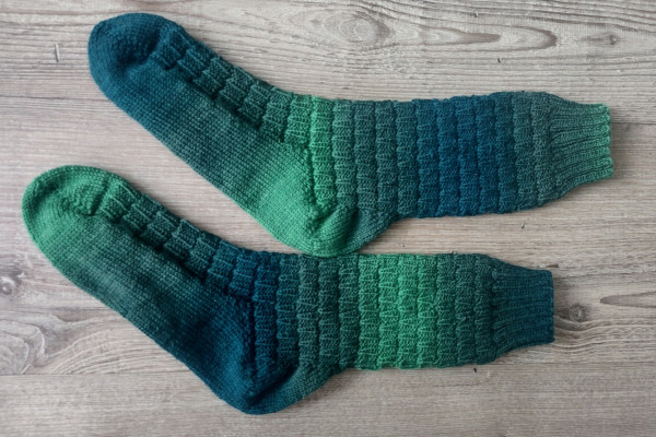 A pair of handknit socks, colors are a gradient of light to dark green. Color sequence on the socks doesn't match. A textured pattern on the leg and instep. 