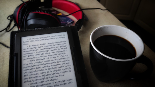 na stole czytnik ebooków, czarny kubek z kawą i czarno-czerwone nauszne słuchawki. 