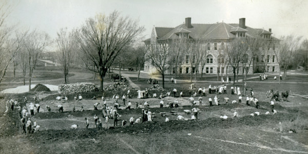 Young men with picks and shovels dig long, rectangular trenches in a lawn. Women in light-colored dresses are observing. A line of wheelbarrows is parked within the boundaries of the foundation. Large stacks of stone are in the background. A three-story stone structure is in the background.