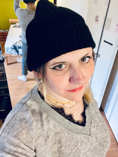 Selfie from a slightly tilting downwards perspective. I’m wearing a black beanie, gray sweater, a necklace with yellow beads and a little chain, as well as light makeup: eyeliner and orange-y shiny lipstick. I am smiling and looking at you in an enticing way. 