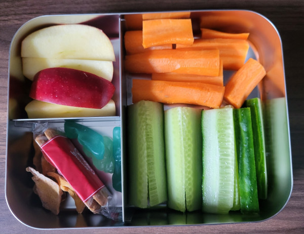 A lunch box with three compartments. One with aplle slices, one with dried mango, lotus cookies and Katjes wine gums. A larger compartment with cucumber and carror sticks.