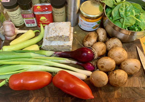 A cutting board on which are placeed: a head of red garlic, 3 yellow peppers (2  jalapeño,1 banana), 2 thick squares of tempeh, two red spring onions, two thick scallions, 2 red San Marzano tomatoes, 9 brown mushrooms, spices — Ancho chili pepper, kala namak, red tin of Spanish smoked paprika, a little jar of ground cumin, a pepper mill, and a jar of tumeric paste. A stainless steel bowl is filled with fresh spinach leaves. 