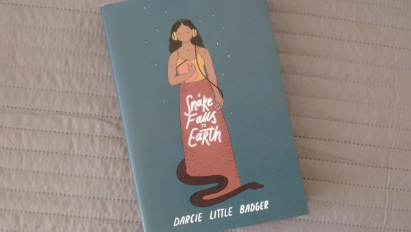 The cover of A Snake Falls to a Earth shows a young woman  --her skin the color of cooked clay, her black hair falling behind her shoulders in soft waves-- who wears a pair of yellow headphones on her head while holding a phone on her hand. She stands, wearing a yellow tank top and a long skirt the color of earth that seems to b made of some woven fabric, against a blue-gray background where fireflies glow. 
A brown snake, a cottonmouth, slithers   from behind her.
The title is written in white letters as it is the name of the author: Darcie Little Badger.