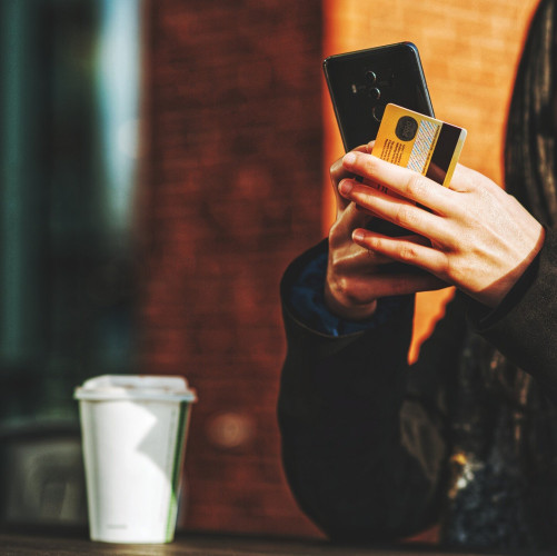 A person's hands holding a phone and a credit card. Elbows propped on a table, a white to-go coffee cup off to the side.