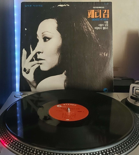 A black vinyl record sits on a turntable. Behind the turntable, a vinyl album outer sleeve is displayed. The front cover shows Patti Kim holding her hand to her face