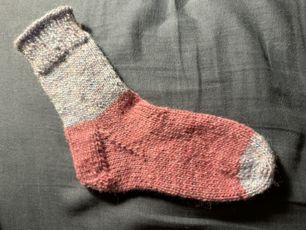 One handknit sock, cuff leg and toe in light blue with speckles of color, heel and foot in pink.
