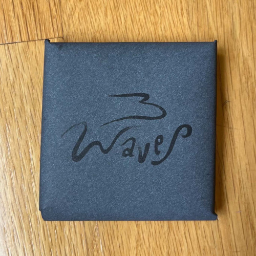 A dark blue, square, hand-folded envelope with the title “Waves” printed in stylized black ink. 