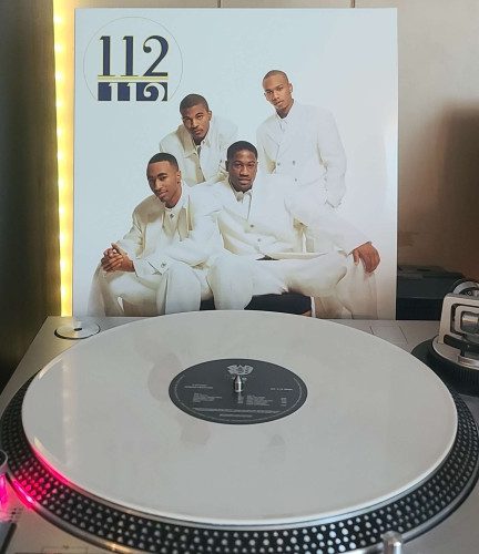 Image shows a turntable with a white vinyl record on the platter. Behind the turntable vinyl album outer sleeve is displayed. The front cover shows the 4 members of 112 looking at the camera in white suits. 2 are standing and 2 are sitting down