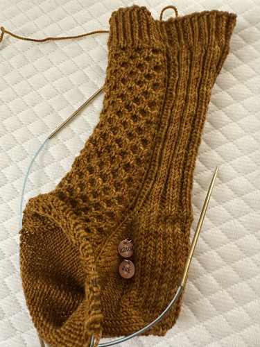 A partially knitted sock on a white background. The sock has a cuff, leg, and heel - but no foot (yet). The yarn is a dark honey color, and there is a honeycomb pattern on the front of the sock. If has two circular needles in the fabric-in-progress. 