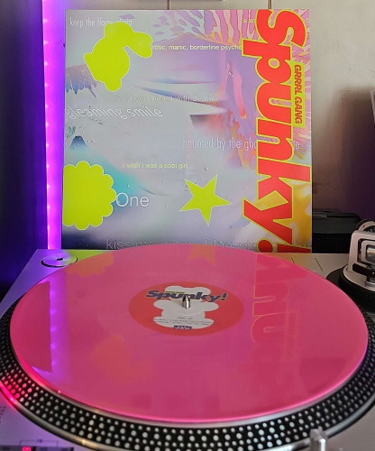 A pink vinyl record sits on a turntable. Behind the turntable, a vinyl album outer sleeve is displayed. The front cover shows artist and album title name, along with random phrases and shapes. 