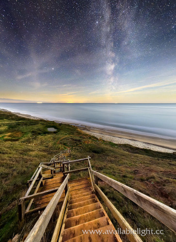 A steep and lengthy tiered set of wooden steps leading down to a deserted beach. The night time sky is full of stars and the Milky Way is visible. The sea is flat calm, scrubland made from a eroding cliff surrounds the steps. The colours are blues, purples, yellow on the horizon. Ships out at sea appear as a yellow glow in the distance. A calming scene with an isolated feel. 
