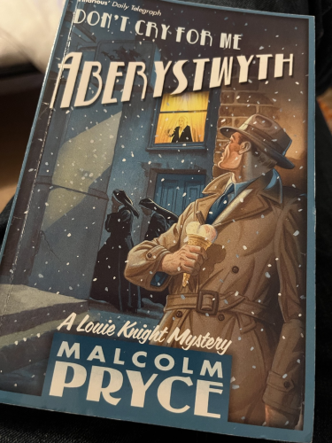 Front cover of Don’t Cry For Me Aberystwyth with Detective Louie Knight in traditional noir outfit of brown raincoat and hat pressed against a wall with ice-cream in hand looking up at a man in a window while two nuns move past in the snow.