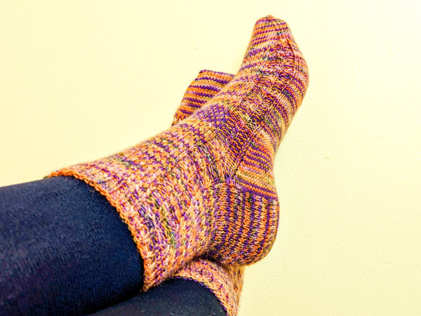 Close up of my feet in a completed pair of hand-knitted socks, worn over thick black tights. The socks are knitted from an orange and purple striped yarn, with a slip stitch pattern breaking up the colour stripes. My left foot is crossed over the right, showing a cable detail that runs down both sides of each sock.