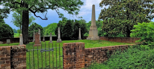 An old graveyard contained behind a brick wall. The gate has a decorative scroll label that reads "Madison." There is a particularly tall obelisk grave marker in the back corner of the walled cemetery, marking James Madison's grave. The grass and trees surrounding the cemetery are a lush green. 