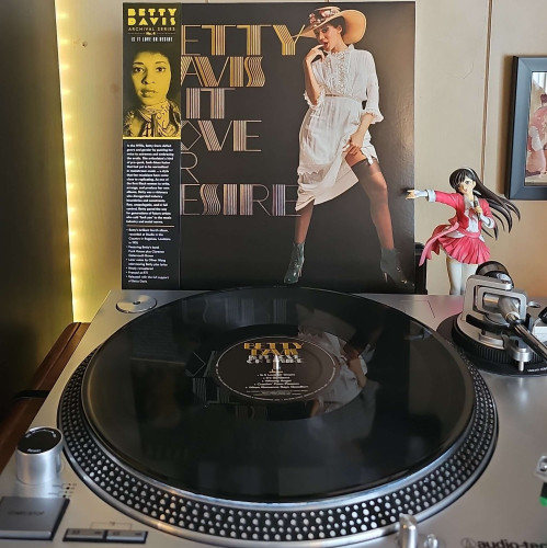 A black vinyl record sits on a turntable. Behind the turntable, a vinyl album outer sleeve is displayed. The front cover shows Betty Davis wearing a white dress and a sun hat. She is lifting up part of the dress to expose her thigh, while biting on her finger seductively. 