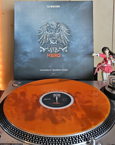 A translucent orange vinyl record sits on a turntable. Behind the turntable, a vinyl album outer sleeve is displayed. The front cover shows Shinhwa's Coat of Arms logo, a banner that says 18th Anniversary, and text that says HERO. 

To the right of the album cover is an anime figure of Yuki Morikawa singing in to a microphone and holding her arm out. 
