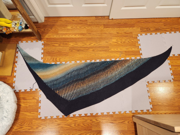A triangular garter stitch and brioche  shawl in dark blue and a variegated yarn in peaches and blues. It is pinned out on blocking mats.