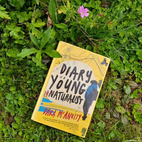 A copy of Diary of a Young Naturalist lying on grass and some other small plants. Above the book is a small pink flower. The cover of the book is yellow with an illustration of a boy with a backpack walking away on a beach