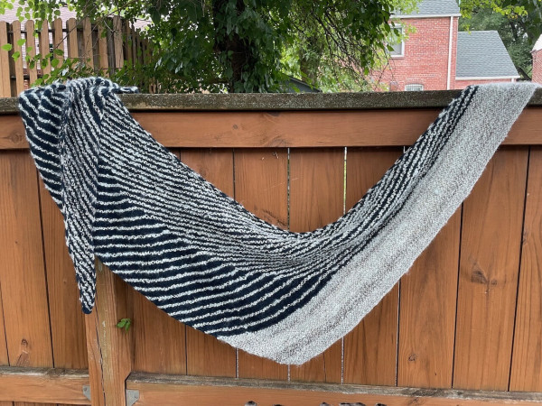 A hand knit shawl of blue and off white yarn hanging on a brown fence in the backyard. The shawl is patterned in stripes of blue and the off white yarn. On the right hand side, the shawl is bordered by several rows of the lighter yarn.
