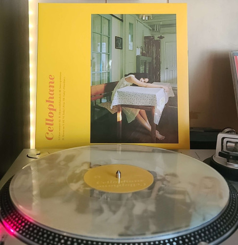 A transparent light yellow vinyl record sits on a turntable. Behind the turntable, a vinyl album outer sleeve is displayed. The front cover shows singer Fromm in a room sitting on a bench, with her chest, arms and head laying across a table