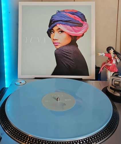 A baby blue vinyl record sits on a turntable. Behind the turntable, a vinyl album outer sleeve is displayed. The front cover shows Yuna sideways turning her head and looking towards the camera. 

To the right of the album cover is an anime figure of Yuki Morikawa singing in to a microphone and holding her arm out. 