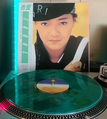 A Transparent Turquoise vinyl record sits on a turntable. Behind the turntable, a vinyl album outer sleeve is displayed. The front cover shows ANRI face shot. She is looking at the camera.