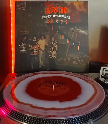 A Red & White swirl vinyl record sits on a turntable. Behind the turntable, a vinyl album outer sleeve is displayed. The front cover shows 4 members of Bone Thugs~N~Harmony standing in a dilapidated inner city with skulls and bones on the ground