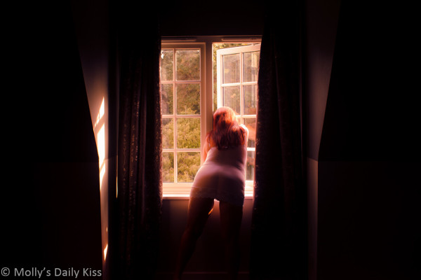 Molly wearing white slip looking out of window. The sinlight is cutting in from the side and illumating her red hair and on the inside of one is her thighs. The rest of the room is in shadows