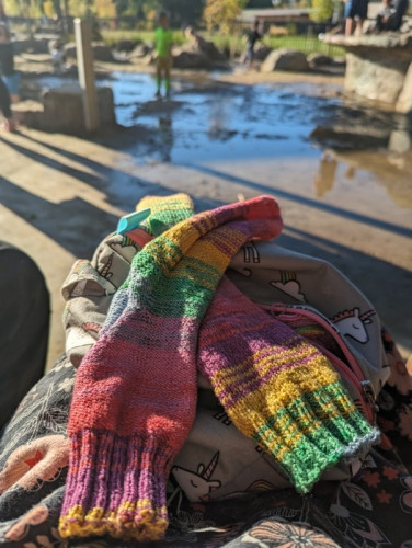 A pair of fairly rumpled looking handknit socks with a bright self striping pattern in a rainbow of colours. In the background is a play area with sand and a lot of water reflecting a bright blue sky 
