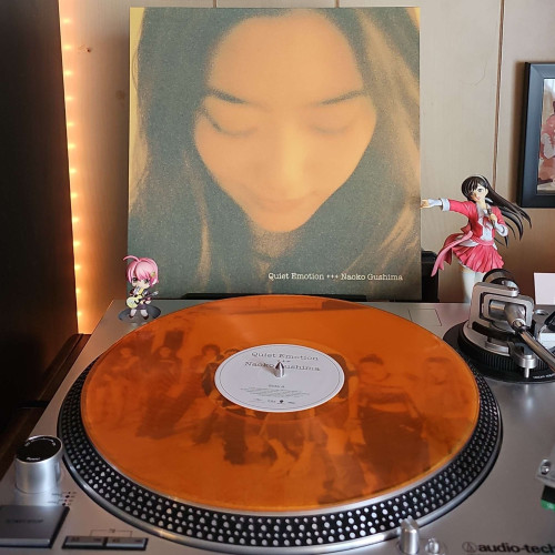 An orange transparent vinyl record sits on a turntable. Behind the turntable, a vinyl album outer sleeve is displayed. The front cover shows Naoko Gushima's headshot. She is looking down. 