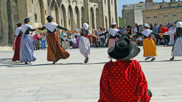 A young boy dressed in a traditional dress watches a group of people dancing the farandole, a traditional dance, in Avignon, France. 

© Adobe Stock – Ph. illustrez-vous