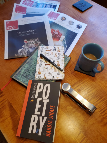The Best Canadian Poetry 2024 collection (guest edited by Bardia Sinaee, Biblioasis) sits on a wooden dining room table with a notebook with an ornate green cover, a notebook with cartoon dogs on the cover, a black pen, a silver stapler, coffee in a blue cup and sections of the Saturday Globe and Mail newspaper
