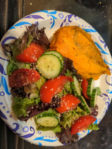 Spring green salad with cucumber, tomato, Asian salad dressing, and hemp seeds, with a peeled baked sweet potato on the side, on a paper plate.