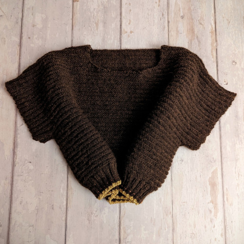 A brown knitting WIP on a white wood-effect background. The knitting is the body of a sweater, with the top seamed together, the shoulders ready to become dropped sleeves, and neck shaping. Most of the body is dark brown, with a pattern of horizontal textured stripes, and a thin gold border at the bottom edge. There is a pink lifeline just up from the border, barely visible.