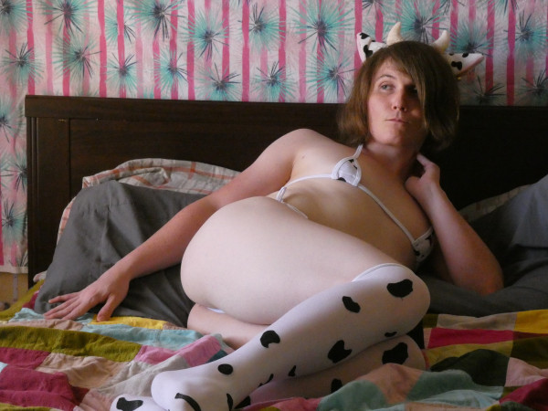 A picture of me laying on my bed with a cow bikini, cow thigh highs, and a cow headband on, staring off into the distance.