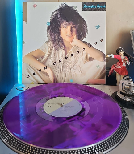 A translucent purple vinyl record sits on a turntable. Behind the turntable, a vinyl album outer sleeve is displayed. The front cover shows Miki Matsubara sitting down with disheveled hair, and smiling while resting her hand on her cheeck. . 

To the right of the album cover is an anime figure of Yuki Morikawa singing in to a microphone and holding her arm out. 